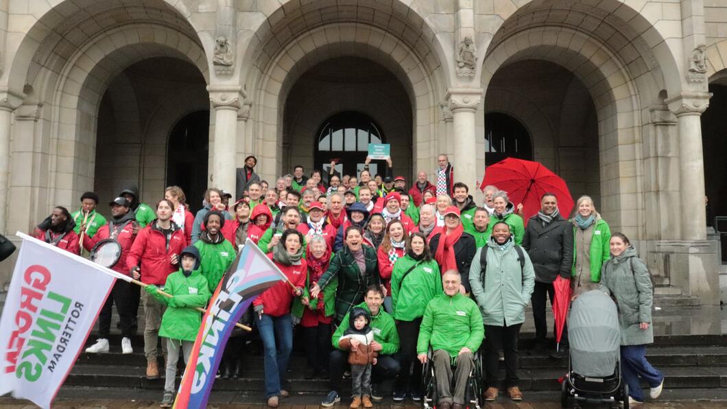 Grote groep vrolijke mensen met GroenLinks-jasjes en PvdA-jasjes staan voor het Rotterdamse Stadhuis