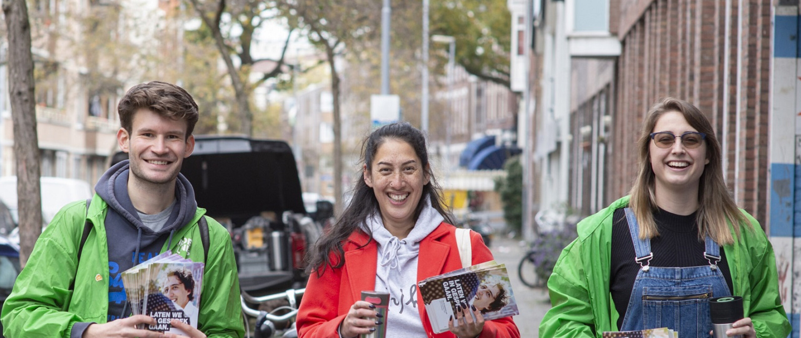 Drie vrijwilligers kijken lachend in de camera tijdens een flyeractie