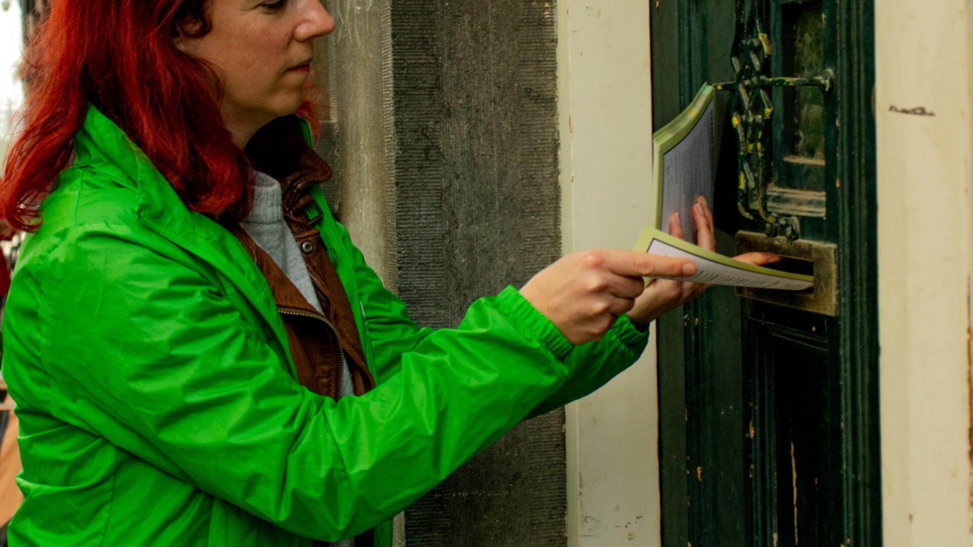 Een persoon met een groene jas doet een flyer door de brievenbus